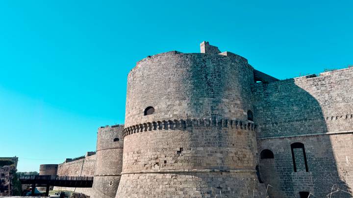 Massive stone towers and walls of a medieval coastal castle reach into a blue sky.