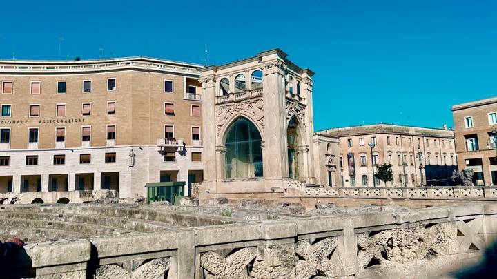 Ancient Roman amphitheater ruins surrounded by 20th-century buildings in a sunny square.