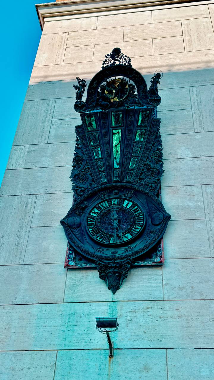 Close-up of an ornate bronze clock with Roman numerals mounted on a stone wall.