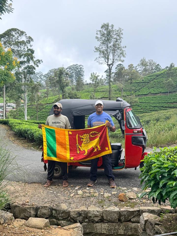 Two men proudly hold the Sri Lankan flag in front of a red tuk-tuk with lush tea plantations behind.