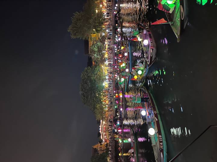 Dozens of tourists ride small wooden boats decorated with colorful lanterns on a river under the night sky, city lights and trees lining the banks.