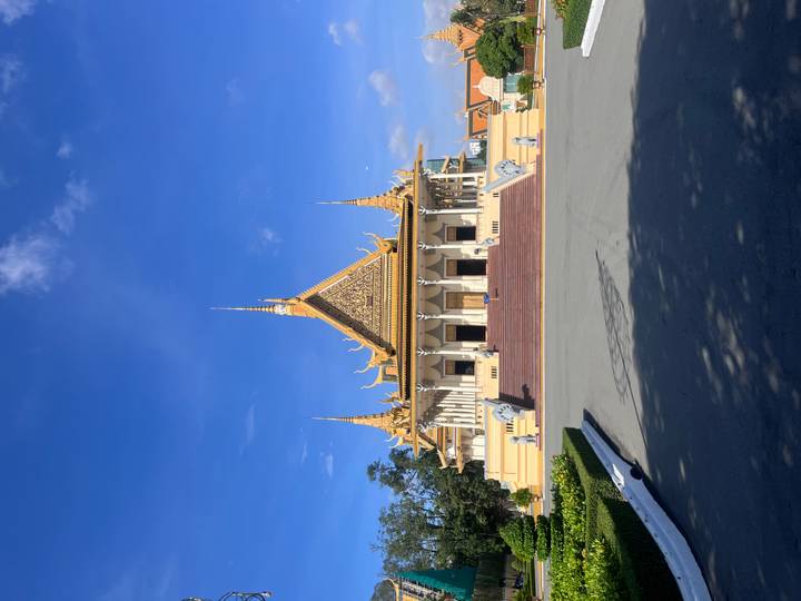 Grand golden-roofed Khmer palace building with ornate spires sits under a clear blue sky, framed by a wide stone courtyard.