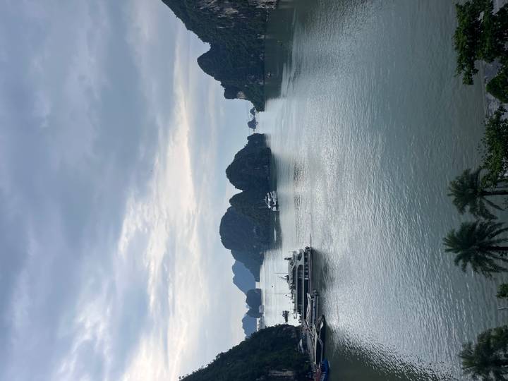 Karst islands rise from calm green waters of a wide bay where cruise boats are anchored under a cloudy sky.
