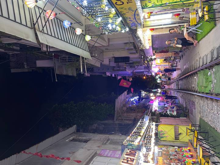 Nightview of Hanoi Train Street with railway tracks flanked by lit cafés, lanterns and people enjoying drinks.