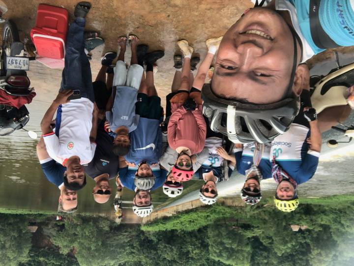 Large group of cyclists pose together on a riverbank with scooters and locals nearby.