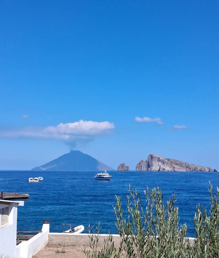 Distant view of steaming Stromboli volcano and rugged offshore islet against clear blue sky.