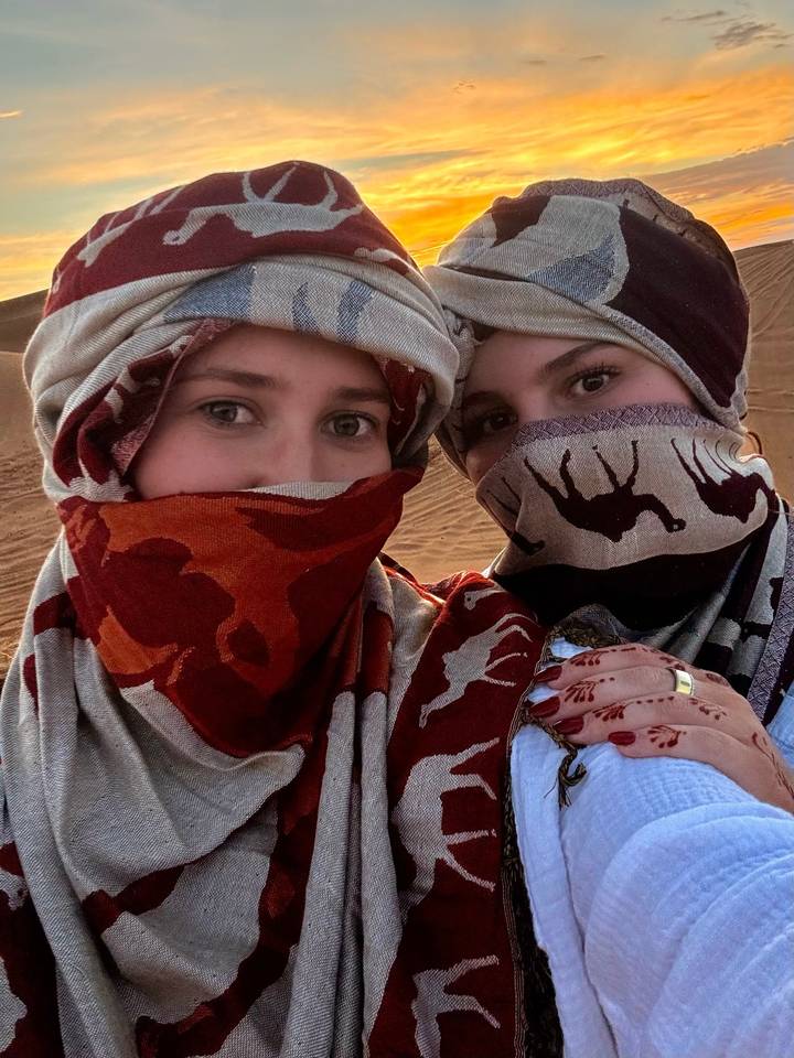 Two women with patterned scarves covering faces take a close-up selfie in the desert.