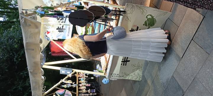 Blonde tourist browses handicraft stalls decorated with wind chimes and fans at an outdoor evening market.