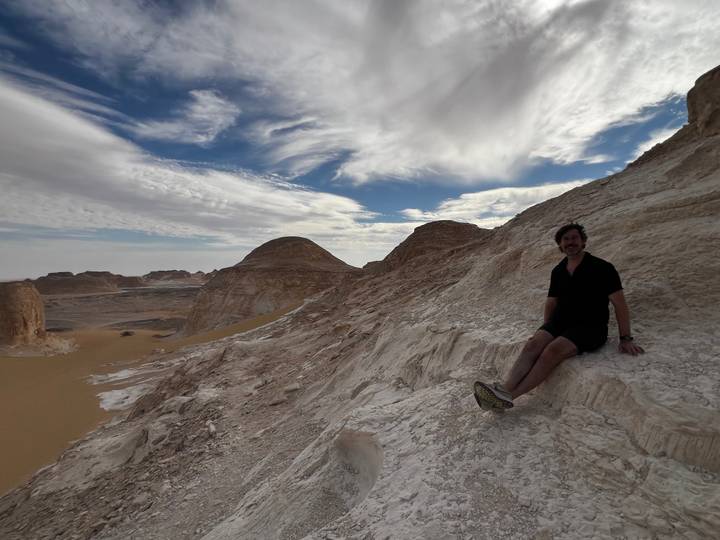 Traveler sits on pale limestone ridge overlooking sweeping desert valley beneath dramatic clouds.