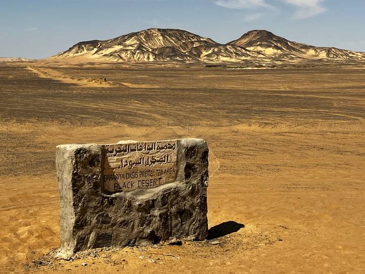 Stone marker engraved with Arabic and English text identifies entry to the Black Desert; striped hills rise in background.