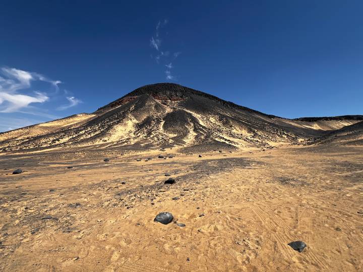 Symmetrical black volcanic hill rises above sandy plain dotted with dark boulders under clear sky.