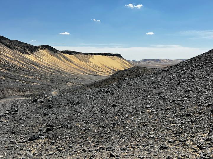 Rugged black volcanic valley with contrasting yellow sand slopes under bright blue sky.