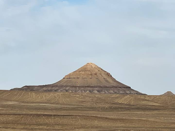 Isolated conical hill rises from flat desert terrain against pale morning sky.