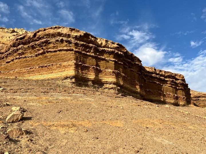 Layered sandstone cliff highlighted by morning sun under blue sky with wispy clouds.