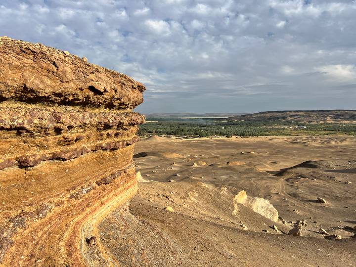 View from layered cliff edge over brown desert plain toward a distant oasis and hills.