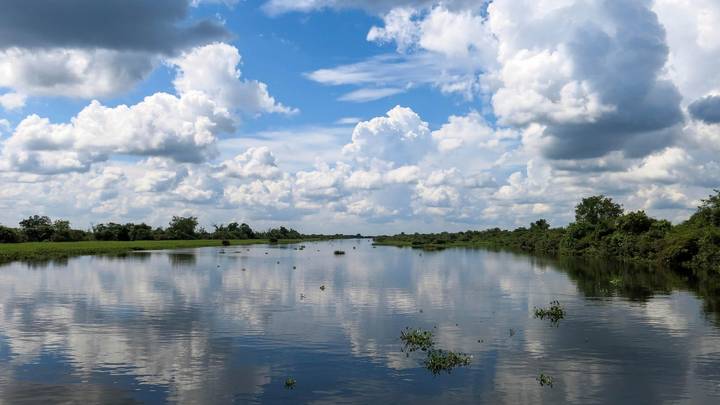 Calm waterway reflecting dramatic cumulus clouds and green banks under a bright blue sky.