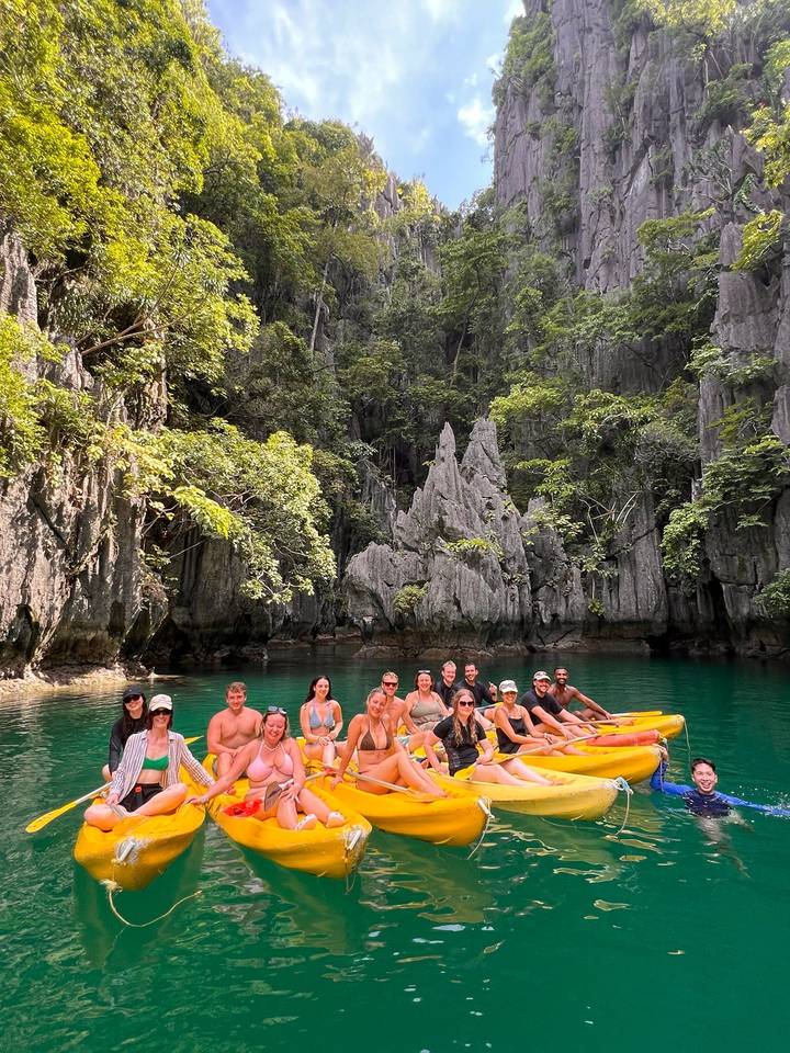 Large group in kayaks floating on emerald water beneath dramatic limestone cliffs and lush jungle.