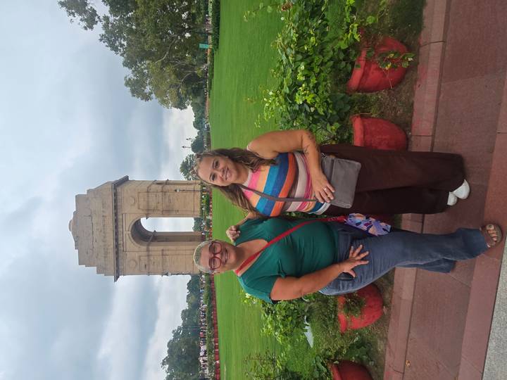 Two women smile for a photo in front of the sandstone India Gate with green lawns around