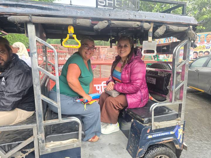 Travellers sit in the back of an open e-rickshaw on a rainy street, wearing casual jackets