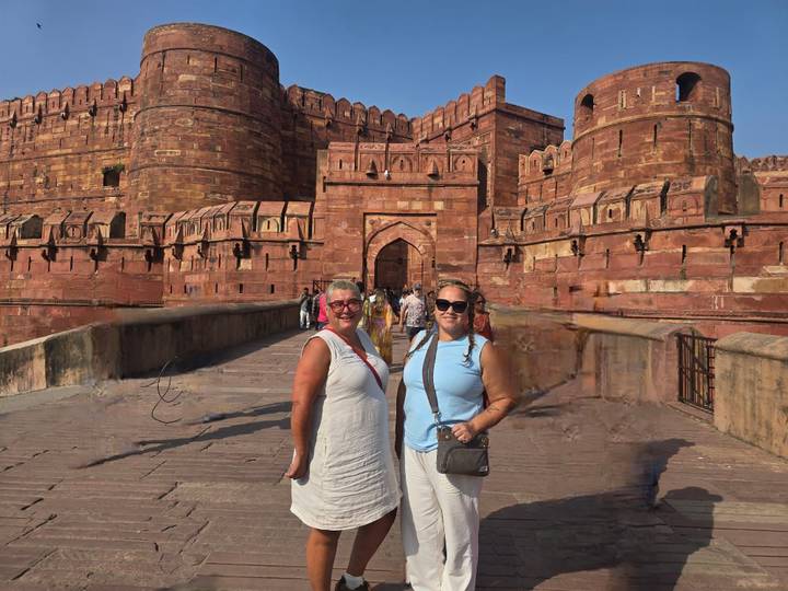 Two travellers pose in front of the red sandstone ramparts of Agra Fort under clear sky