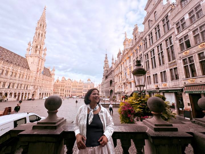 Smiling woman posing in historic European square bordered by ornate Gothic buildings under a cloudy sky.