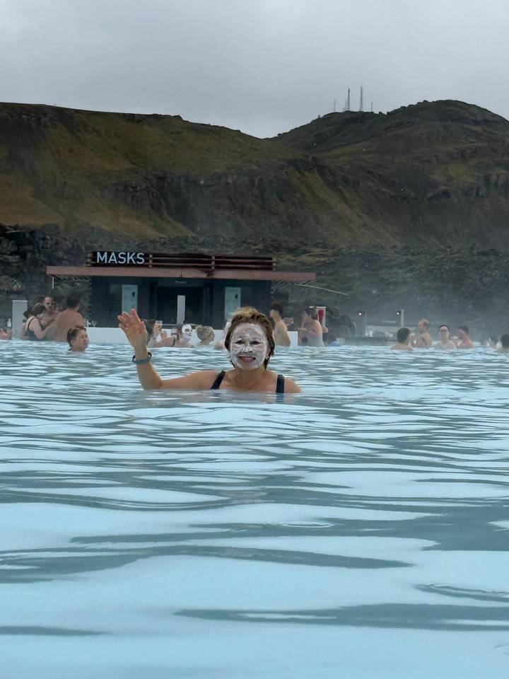 Visitors bathing in milky blue geothermal waters of the Blue Lagoon, one woman waving with silica mask on face.