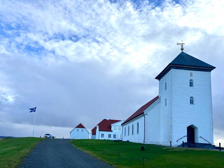 White church complex with red roofs and Icelandic flag set against a vast cloudy sky.