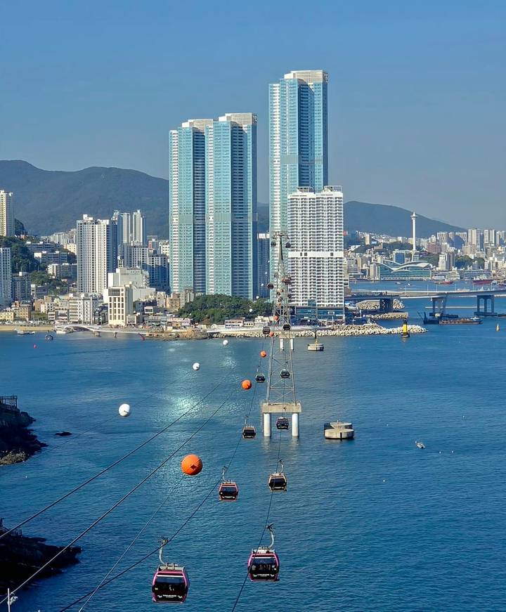 Coastal skyline of Busan with tall towers, cable car crossing the bay and surrounding mountains.