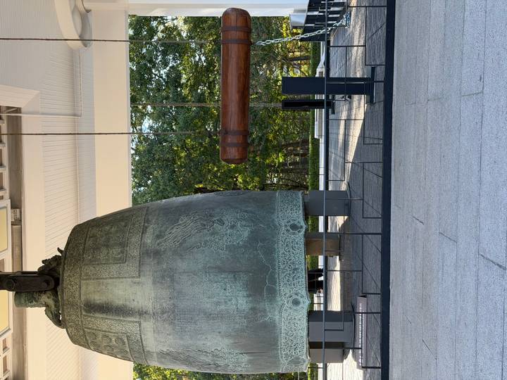 Massive bronze temple bell and wooden striker displayed in an open pavilion surrounded by trees.