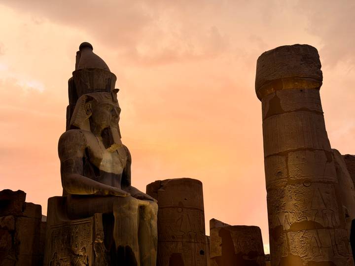 Seated pharaonic statue and towering column silhouetted against a dramatic pink-orange sunset sky in Luxor Temple.