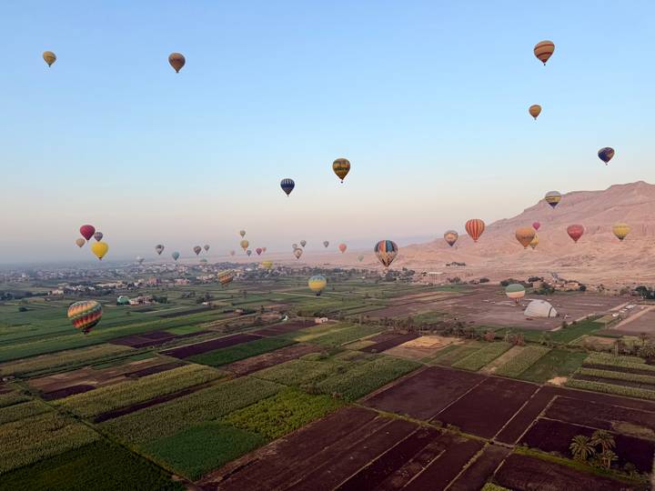 Dozens of colourful hot-air balloons drifting over patchwork green fields and desert hills at dawn
