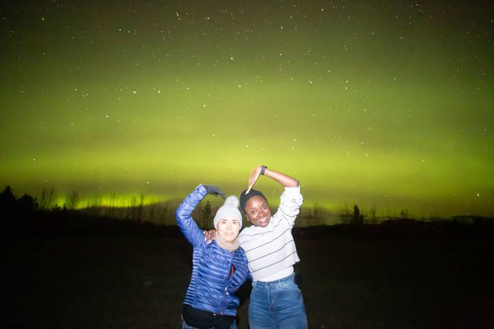 Two friends pose joyfully under a vivid green aurora borealis night sky