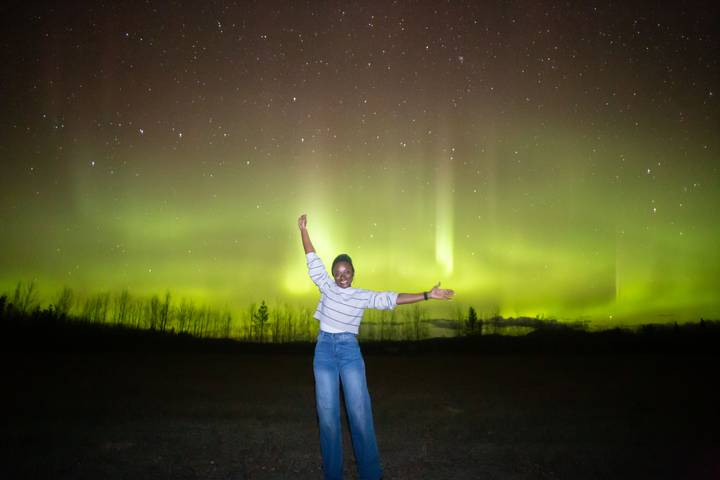 Woman joyfully stretching arms beneath bright curtains of northern lights and starry sky