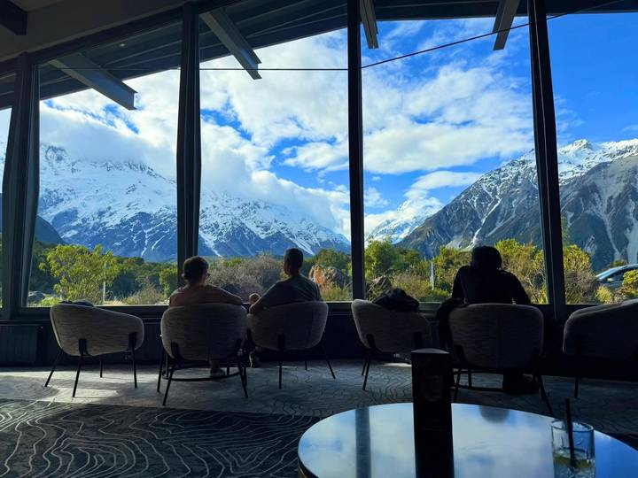 Travelers relax in lounge chairs gazing through large windows at snow-covered mountains and dramatic skies