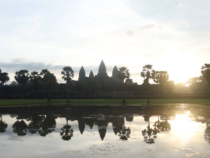 Silhouetted spires of Angkor Wat emerge against a hazy sunrise, reflected in a lotus pond.