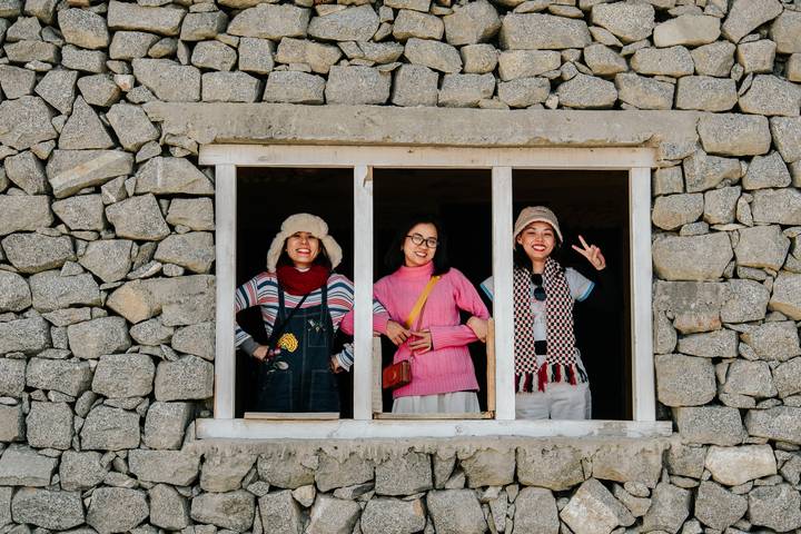Three women smile through a stone-framed window of a traditional mountain house.