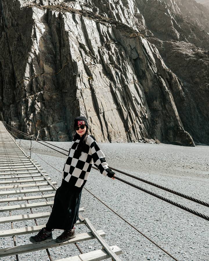 A woman poses on the narrow wooden planks of the Hussaini Suspension Bridge spanning a rocky gorge.
