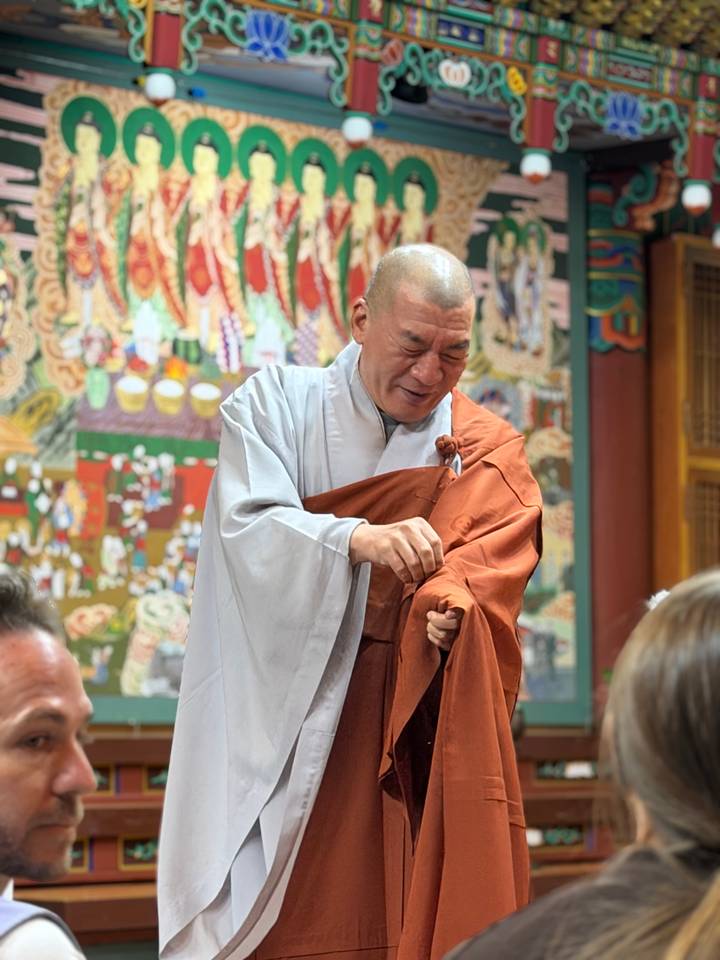 Smiling monk in grey and orange robes prepares a ceremony in a decorated temple hall.