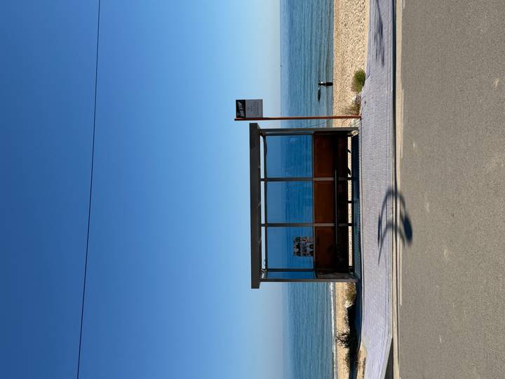 Minimalist coastal bus stop facing a calm blue sea under a clear sky.