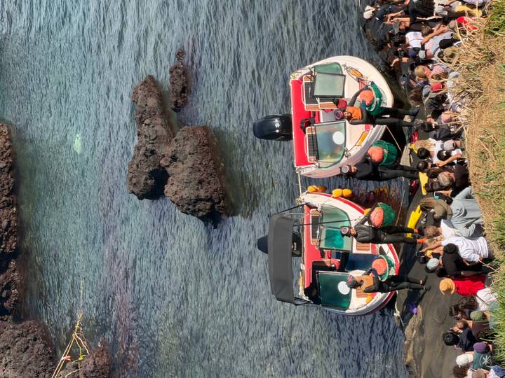 Crowd watching haenyeo divers beside two motorboats docked along rocky Jeju shoreline.