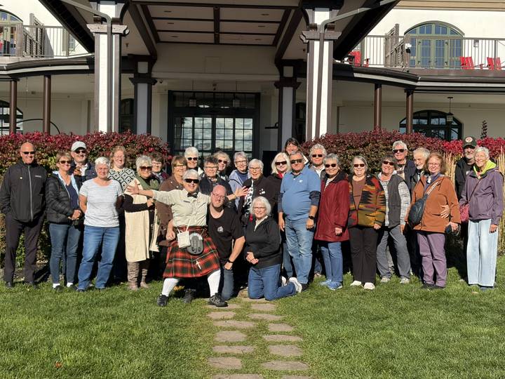 Large tour group poses smiling on a lawn in front of a historic building on a sunny day.