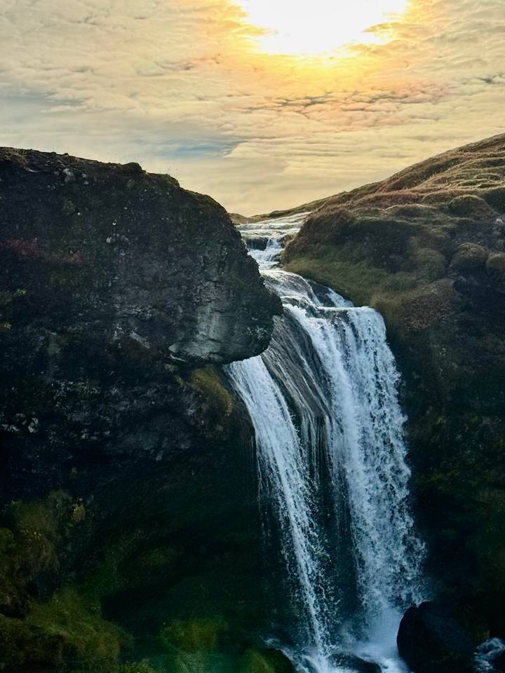 A narrow waterfall cascades between dark volcanic cliffs into a rocky pool at dusk.