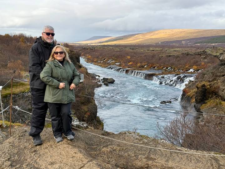 An older couple poses on a cliffside trail overlooking the multi-tiered Hraunfossar waterfall and blue river.