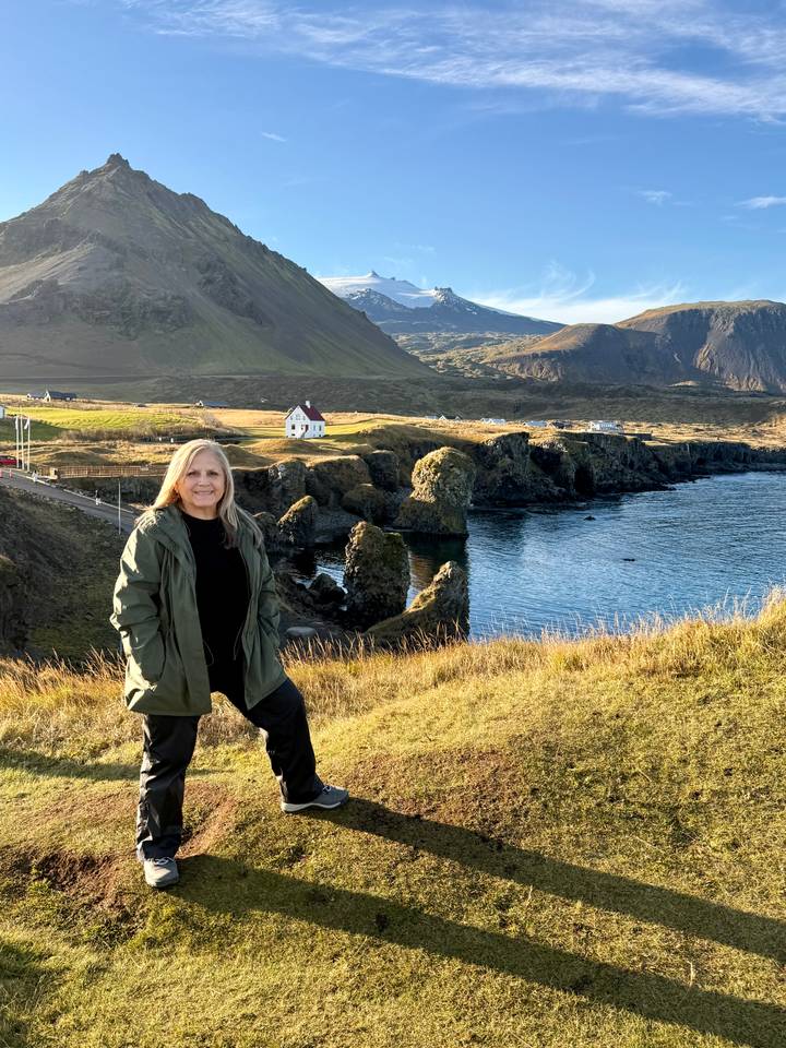 A woman stands on coastal grassland above dramatic basalt sea stacks and a calm blue bay.