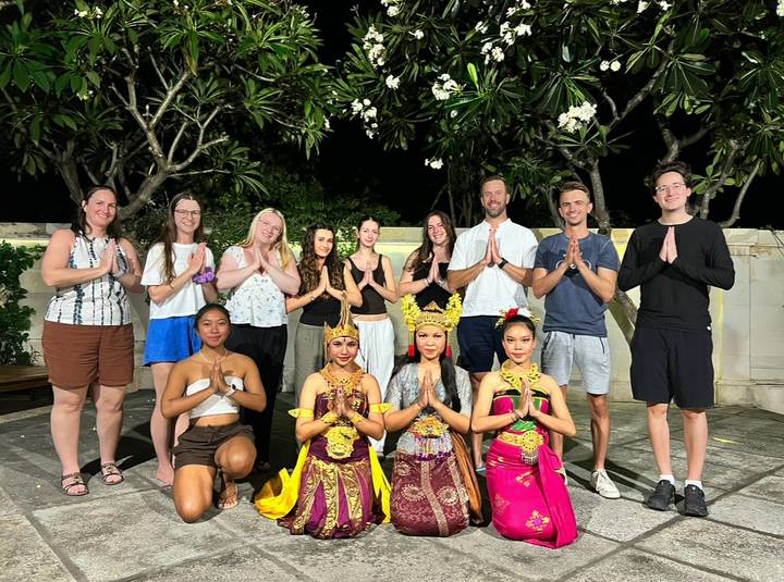 Large tour group posing with Balinese traditional dancers under frangipani trees at night.