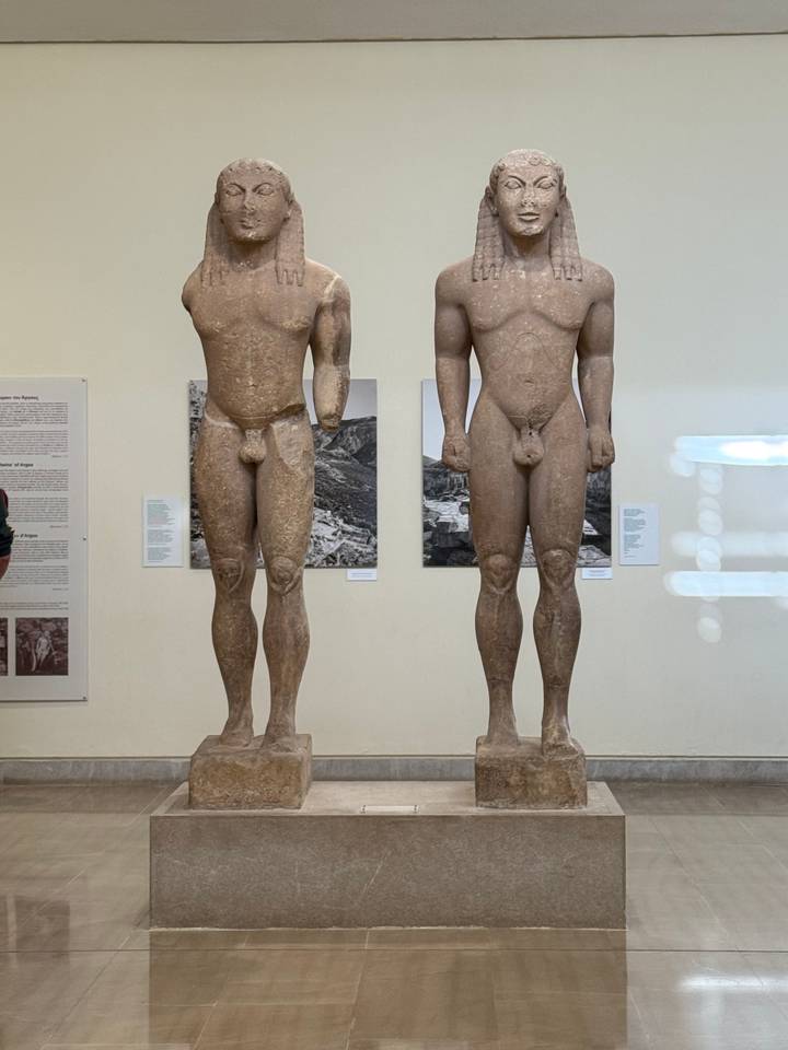 Two ancient kouroi statues displayed in a museum gallery with informational panels behind.