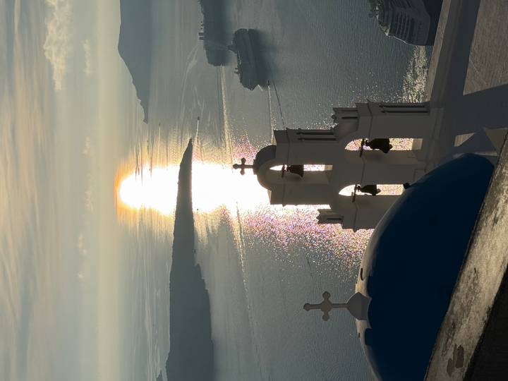 Iconic white bell tower and blue dome of Santorini overlooking the shimmering Aegean Sea at sunset.