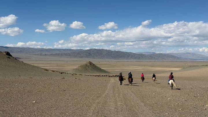 Riders on horseback traverse vast open steppe toward distant Altai mountains under scattered clouds.