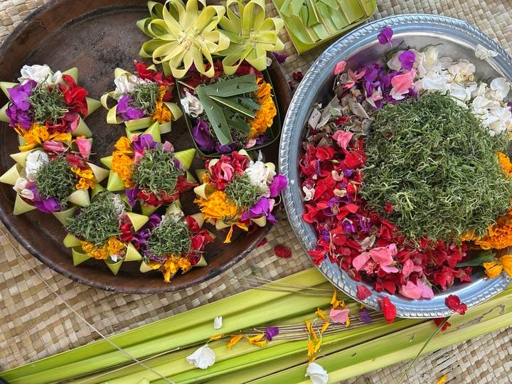 Colourful Balinese flower offerings and petals arranged in wooden and metal trays on a woven mat.