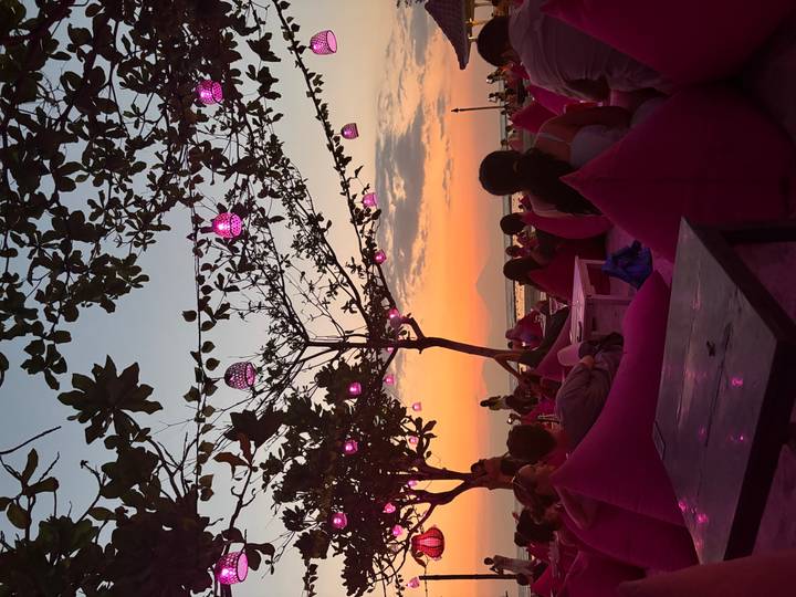 Outdoor beach bar at sunset with pink lanterns hanging from trees, bean-bag seating and relaxed patrons watching the horizon.
